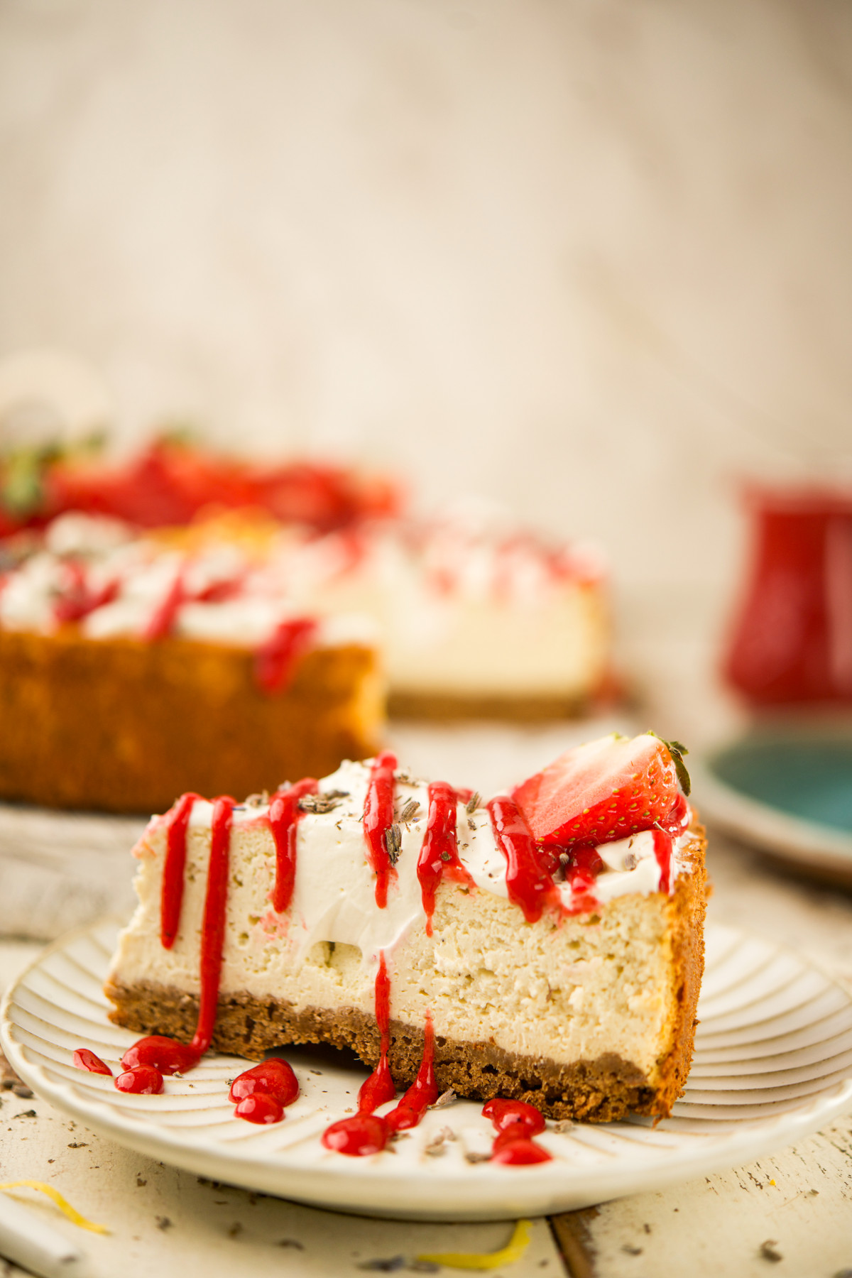 A slice of cheesecake topped with strawberries and red sauce sits on a plate. The rest of the cheesecake is in the background, blurred, with more red sauce and strawberries visible.