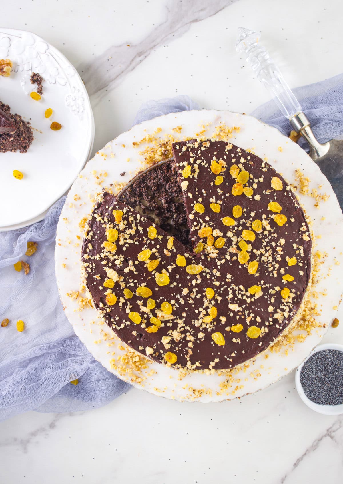 A round chocolate cake topped with yellow raisins and crushed nuts sits on a marble surface, with one slice cut and placed on a white plate nearby. A cake server and a small bowl of seeds are also present.