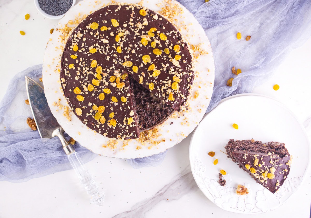 A round poppy seed cake topped with golden raisins and crumbs sits on a white plate. A slice is served on a small plate nearby, with a cake server and a bowl of poppy seeds beside them on a light surface.
