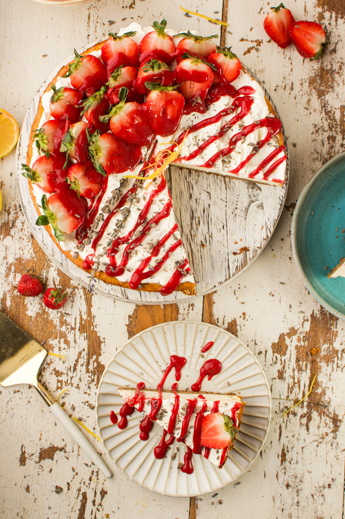 A Strawberry Lavender Cheesecake topped dessert with whipped cream and red sauce is on a rustic wooden table; a slice is cut and served on a plate, garnished with strawberries and sauce.