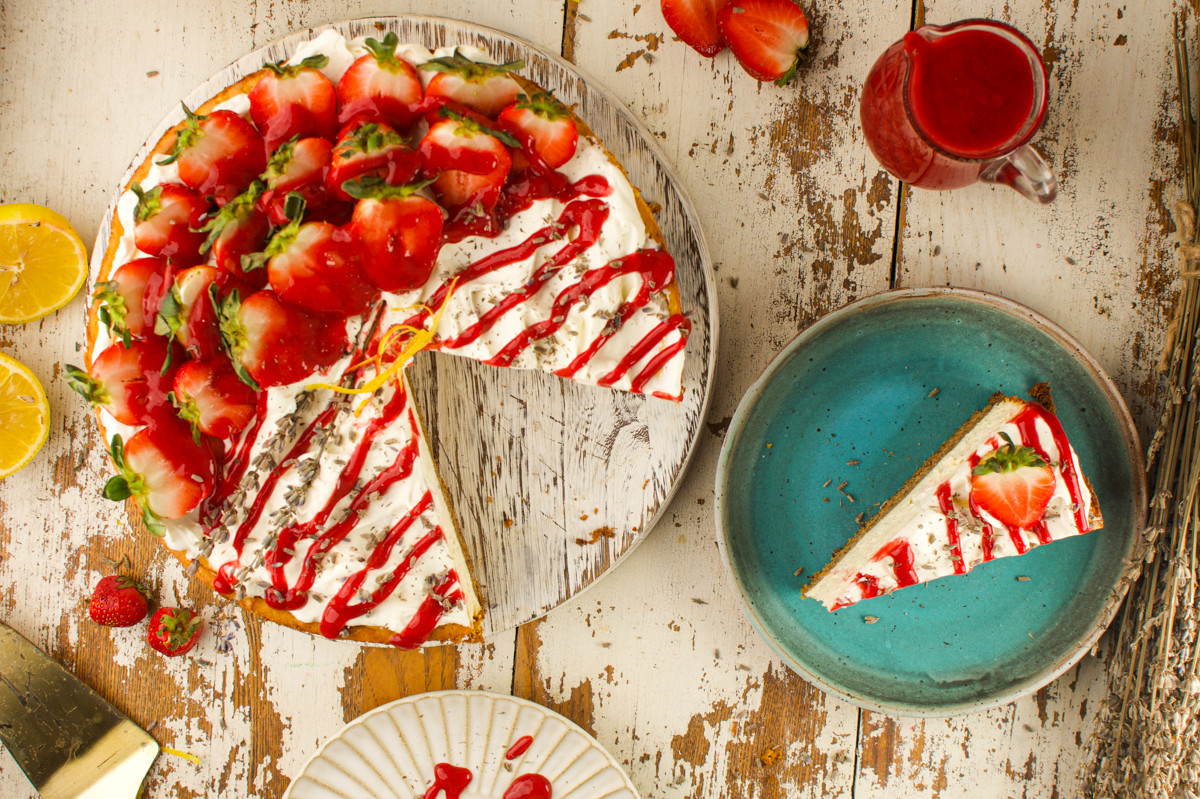 A cheesecake topped with fresh strawberries and red sauce sits on a rustic wooden table. One slice is served on a blue plate, surrounded by a jug of sauce, lemon halves, and scattered strawberries.