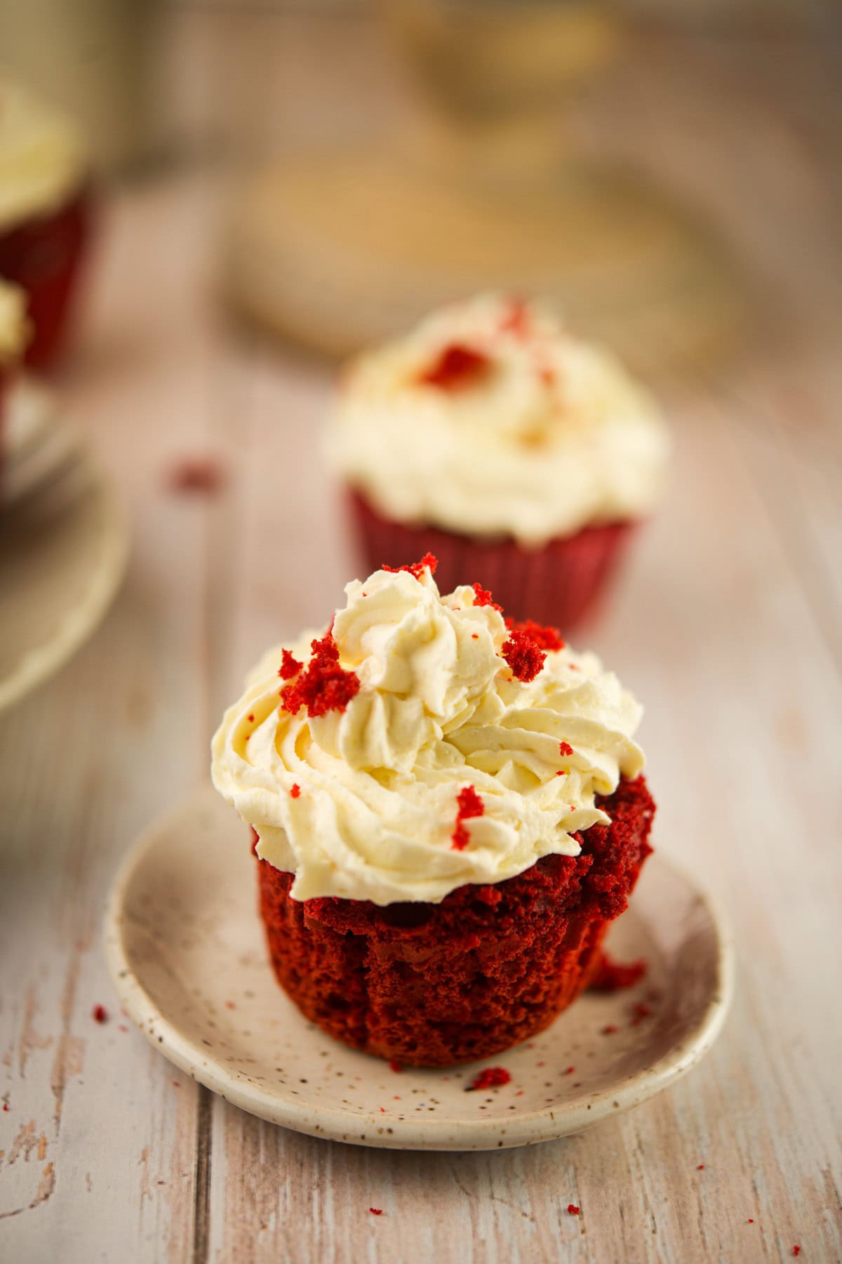 A red velvet cupcake with white cream cheese frosting and red crumbs on top sits on a small plate, with more cupcakes blurred in the background on a wooden surface.