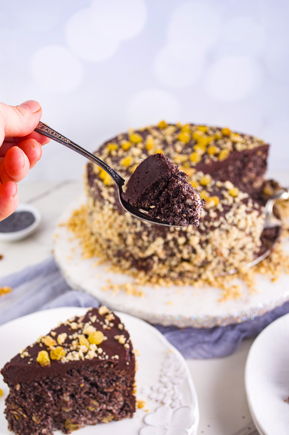 A hand holds up a spoonful of moist chocolate cake with chopped nuts, with the rest of the round cake and a slice on a plate in the background. The cake is topped with chocolate glaze and sprinkled with chopped nuts.