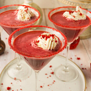 Three martini glasses filled with Creamy Red Velvet Martini Cocktail, topped with whipped cream and red sprinkles. The glass rims are coated with red sugar. Cupcakes and bottles are in the blurred background.