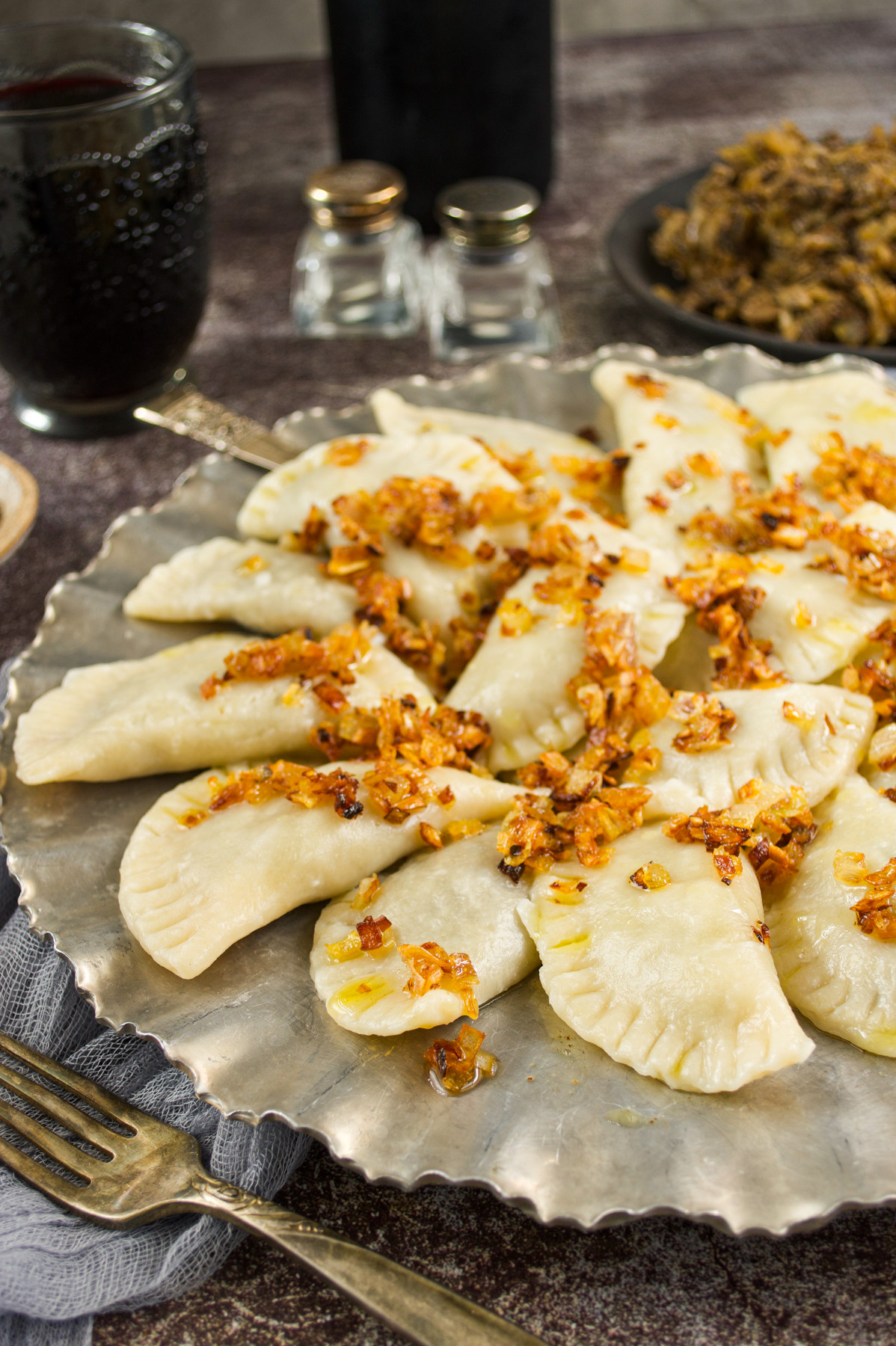 A plate of polish pierogi topped with sautéed onions sits on a textured metal platter, with a fork and knife beside it. In the background, there is a glass of dark drink, salt and pepper shakers, and a dish of cooked filling.