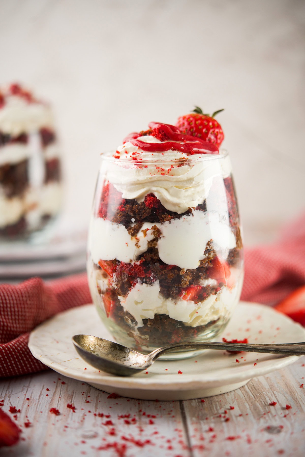 A glass filled with Grain-Free Red Velvet Parfait and strawberries, topped with whipped cream and a strawberry. A spoon rests on the plate beside the glass, with a red cloth and another dessert in the background.
