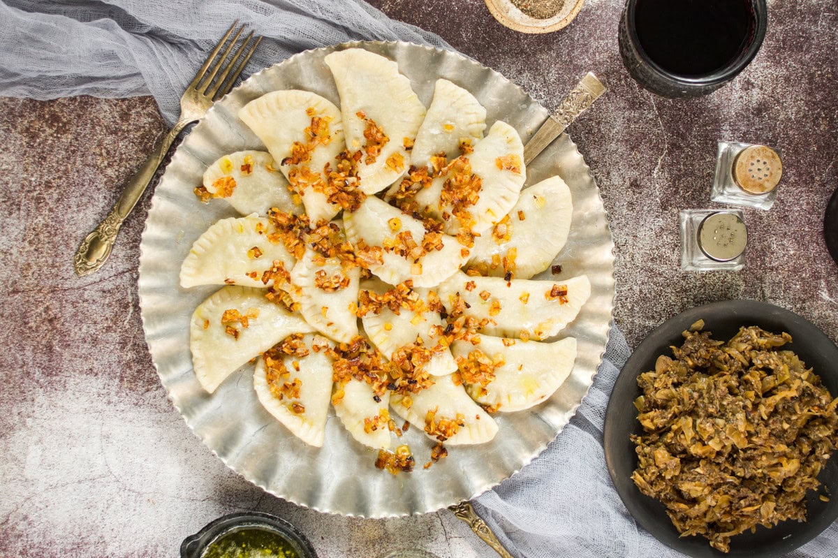 A silver plate filled with crescent-shaped dumplings topped with sautéed onions sits on a table, surrounded by a fork, spices, a glass of dark liquid, and a bowl of stewed vegetables.