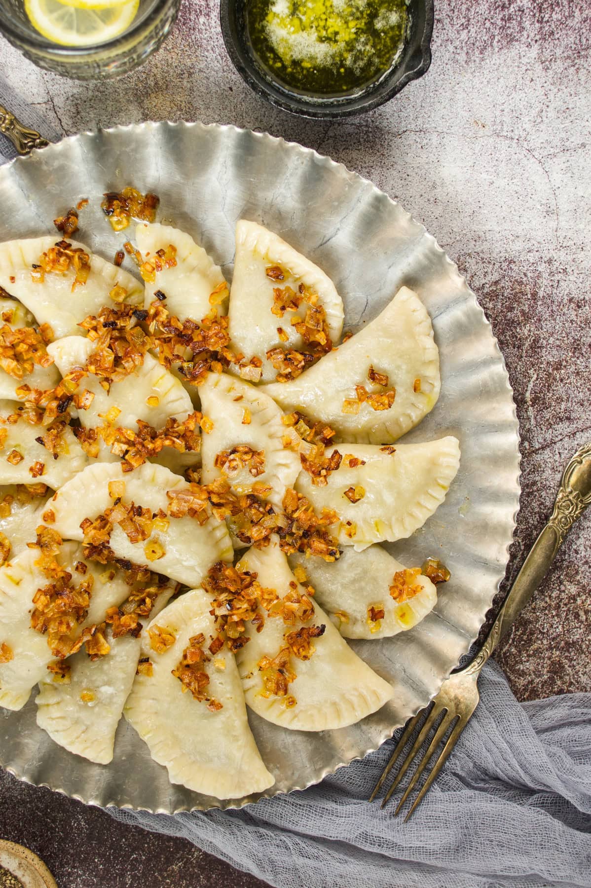A silver plate filled with semicircular dumplings topped with sautéed diced onions, surrounded by utensils, a bowl of melted butter, and a lemon beverage on a textured surface.