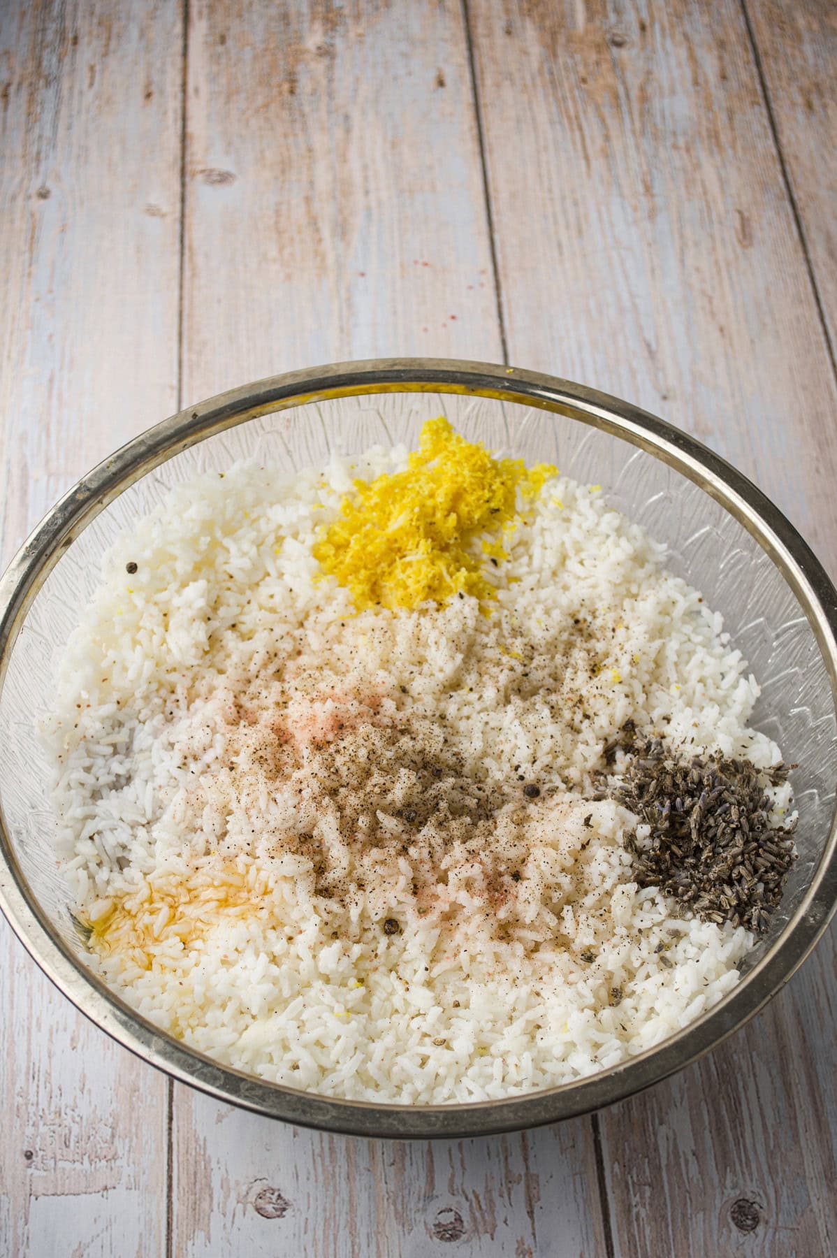 A large glass bowl filled with cooked white rice, topped with piles of black pepper, yellow lemon zest, dried herbs, and a dusting of salt, sitting on a rustic wooden surface.