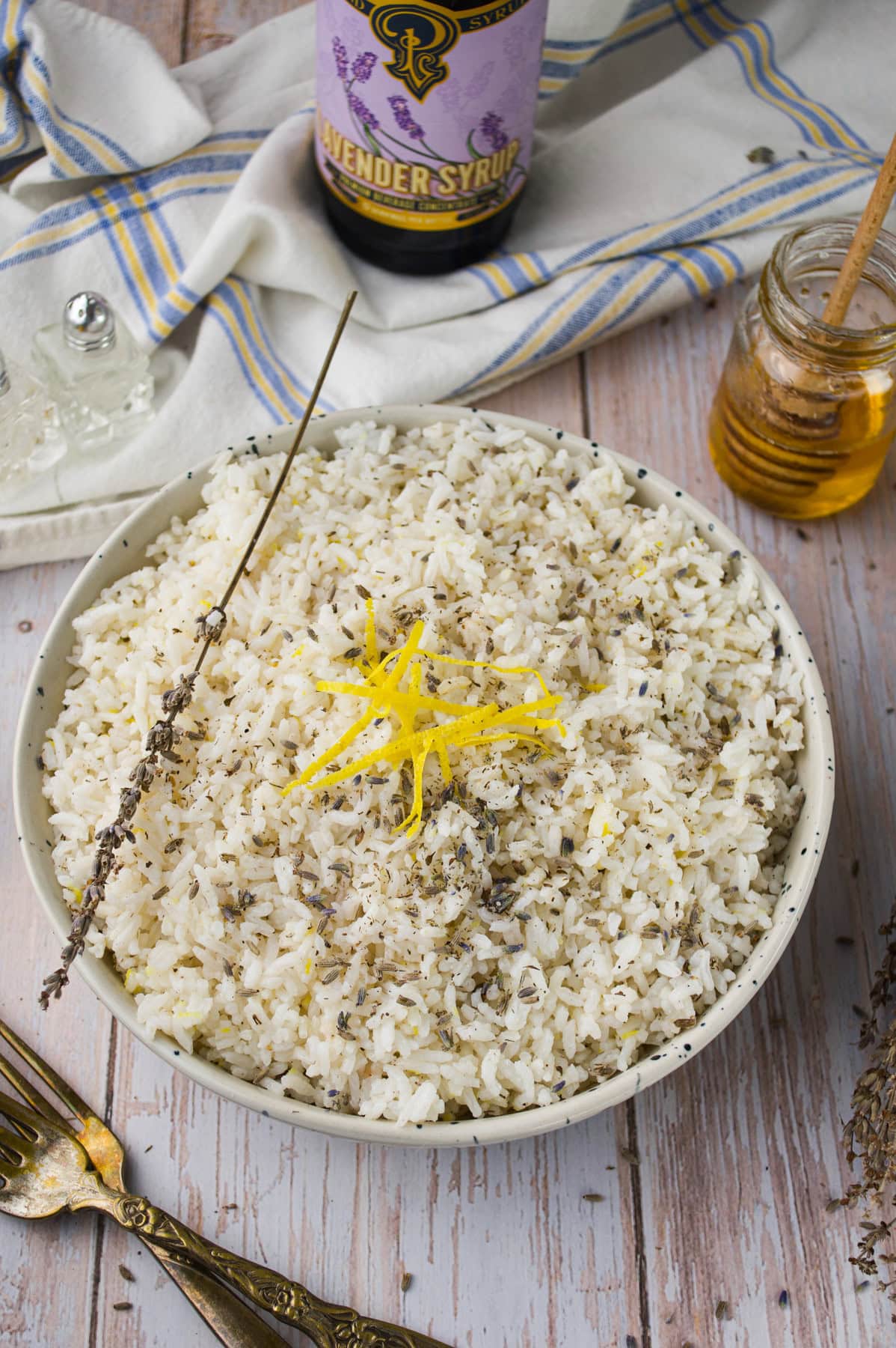 A bowl of white rice garnished with dried herbs and lemon zest, with a sprig of lavender on top. Nearby are a jar of honey, a bottle of lavender syrup, a fork, and a striped cloth napkin on a wooden table.