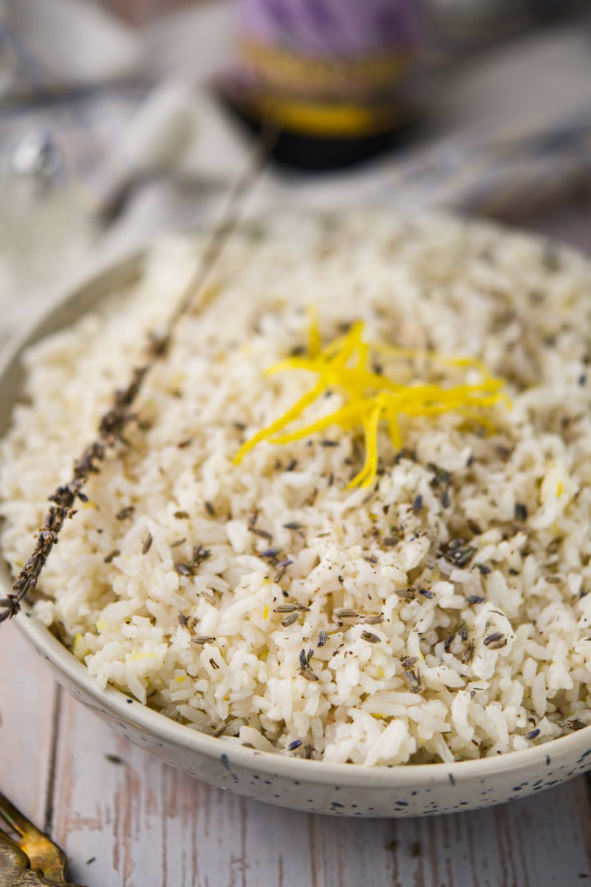 A bowl of Lavender Lemon Rice topped with dried herbs and garnished with yellow lemon zest, with a sprig of dried herbs resting on top. The bowl sits on a wooden surface.