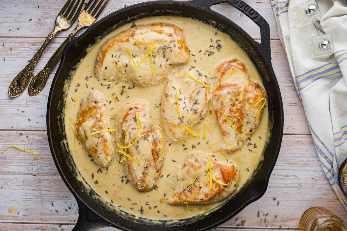 A cast iron skillet filled with cooked chicken breasts in a creamy sauce, garnished with lemon zest and herbs, sits on a light wooden surface near a striped kitchen towel and silverware.