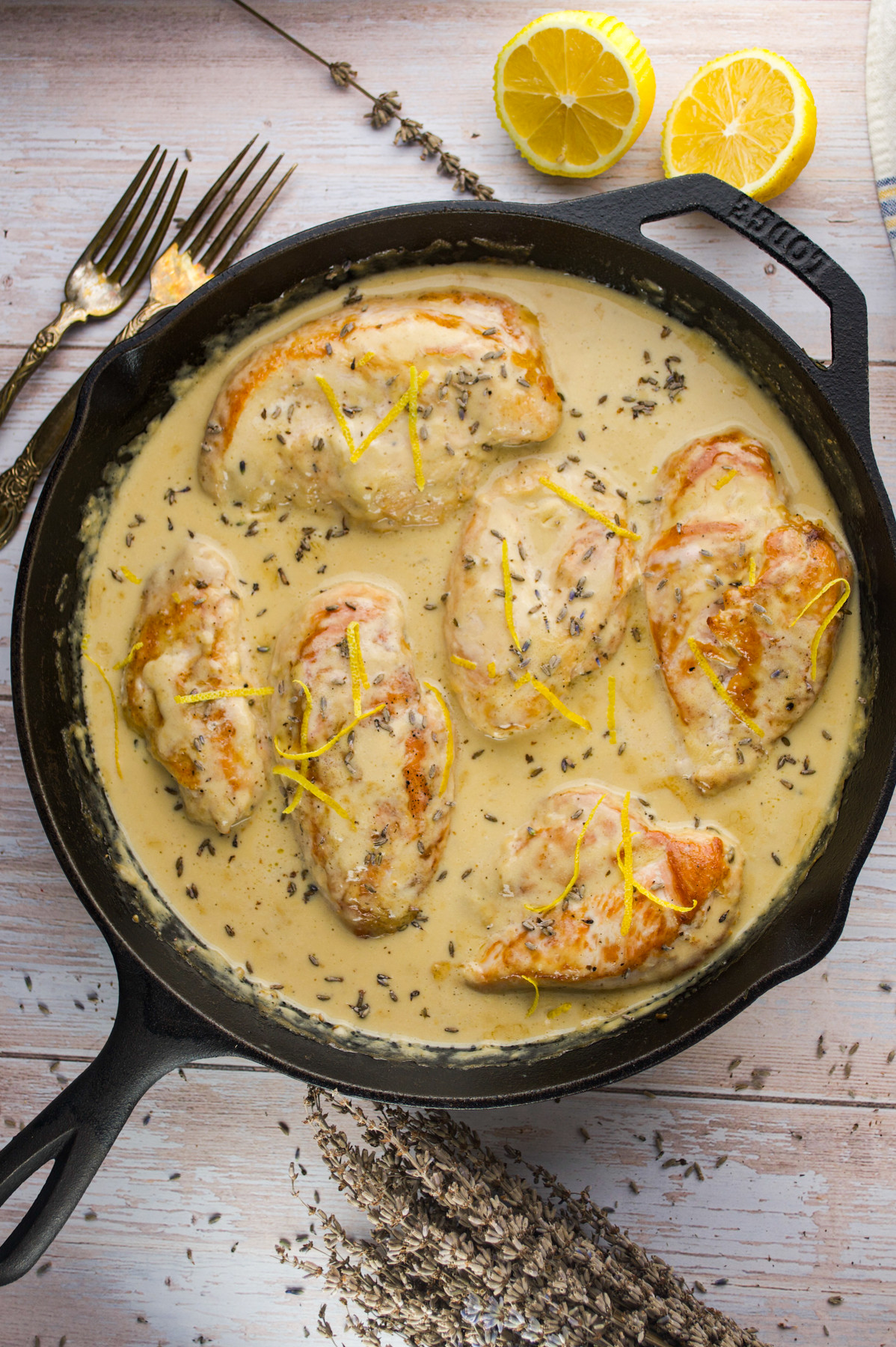 A cast-iron skillet filled with cooked Creamy Lemon Lavender Chicken garnished with lemon zest and dried herbs. Sliced lemons and two forks are placed nearby on a light wooden surface.