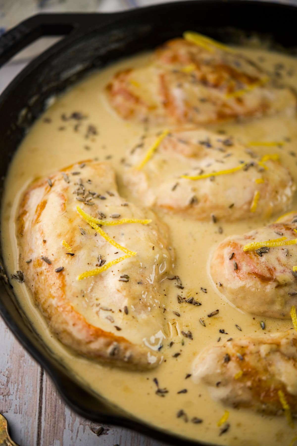 Chicken breasts simmer in a creamy, light brown sauce with visible herbs and lemon zest in a black skillet. The sauce coats the chicken evenly, creating a savory, appetizing dish.