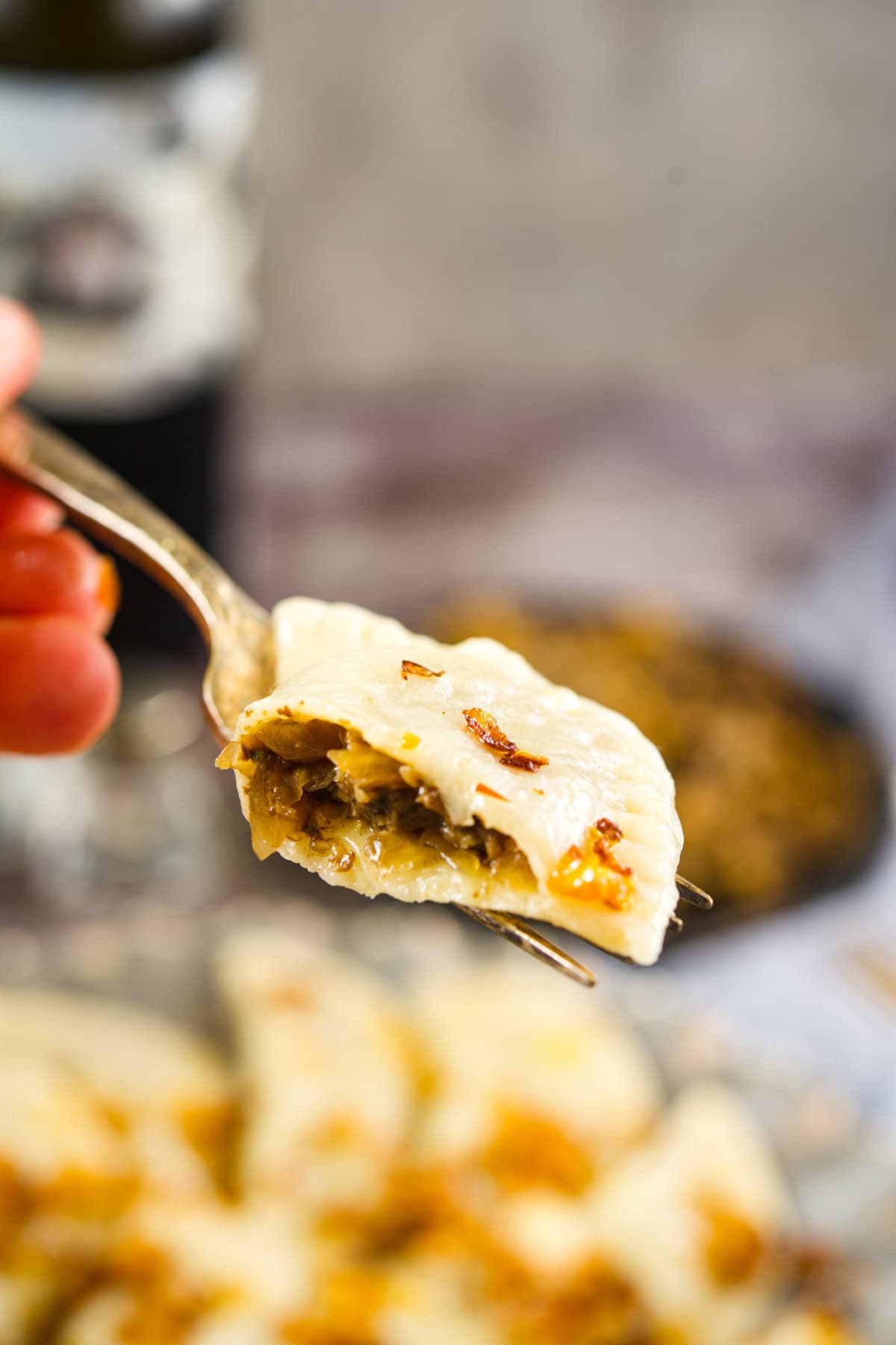 A close-up of a fork holding a half-eaten pierogi filled with a savory mixture, with crispy browned onions on top. Blurred background shows a plate of more pierogi and a dark drink.