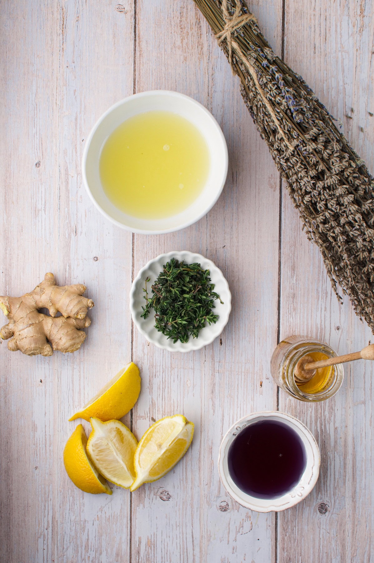 A flat lay of ingredients on a wooden surface: a bowl of pale yellow liquid, a bunch of dried herbs, fresh ginger root, a small dish of fresh herbs, lemon wedges, a small bowl of honey with a dipper, and a bowl of dark liquid.