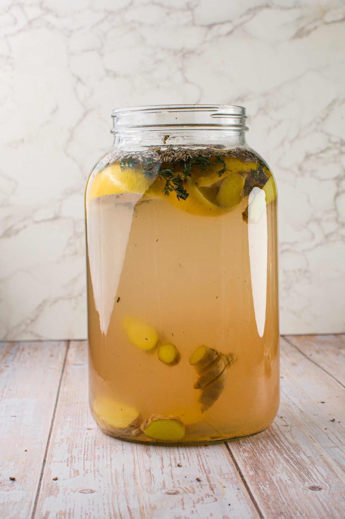 A large glass jar filled with cloudy, light brown liquid containing lemon slices, ginger pieces, and sprigs of herbs, sitting on a wooden surface against a light marble background.