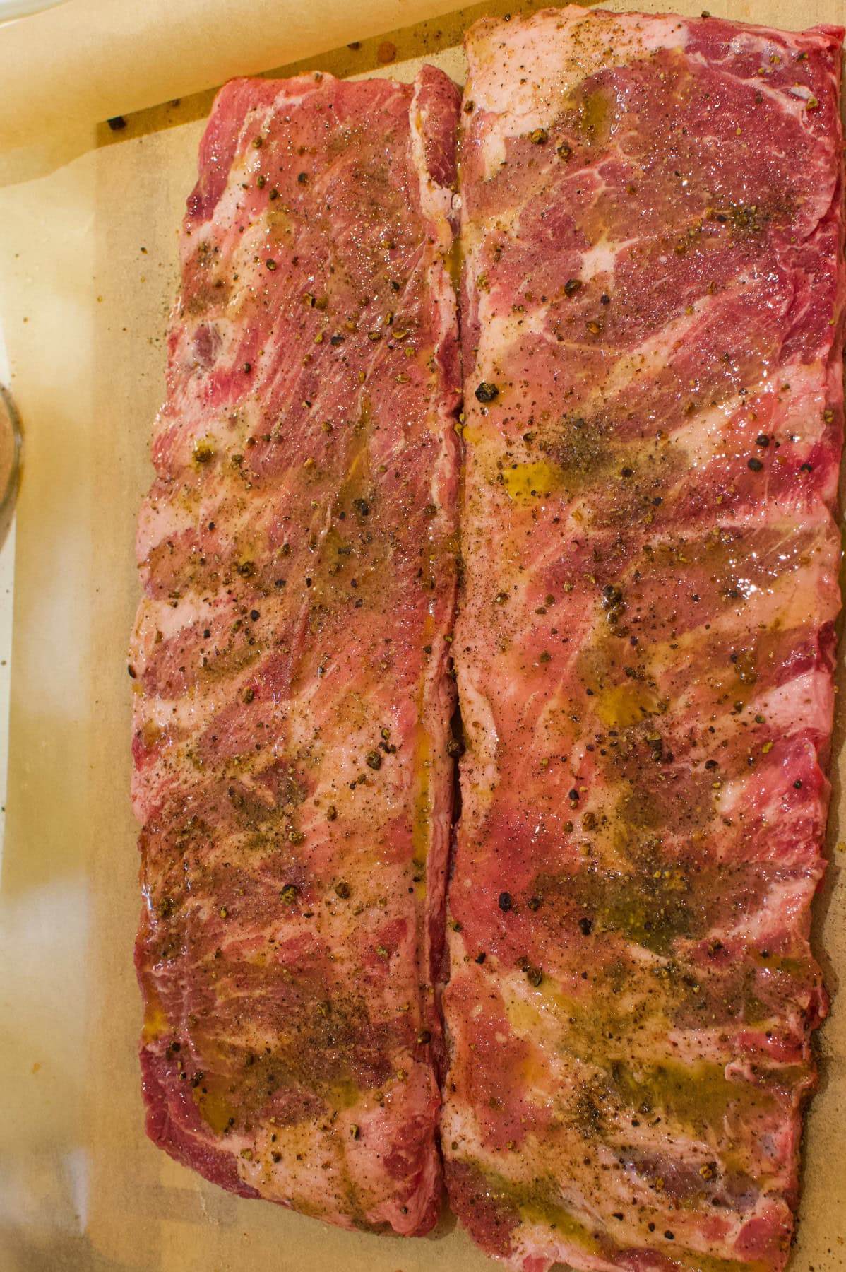 Two racks of raw pork ribs on parchment paper, seasoned with pepper, salt, and drizzled with oil, ready to be cooked.