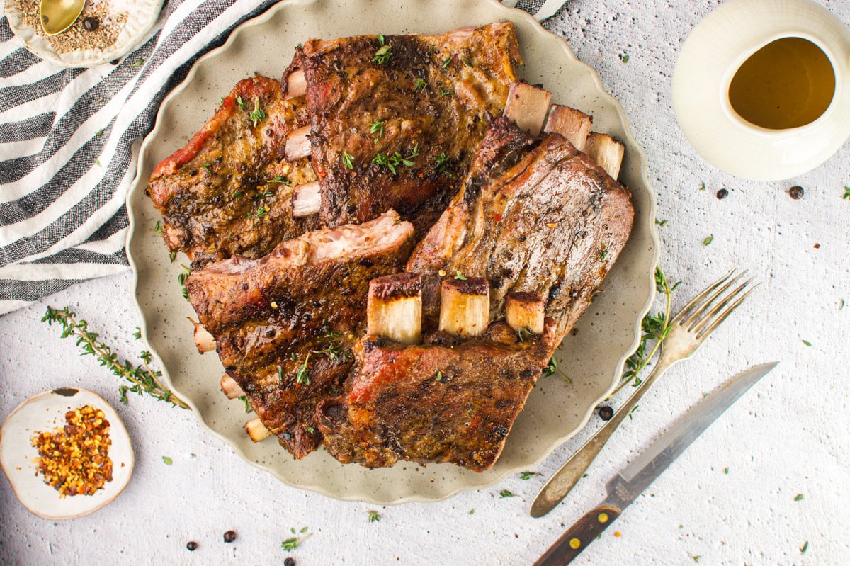A plate of seasoned, cooked beef ribs garnished with herbs, served on a beige plate with a knife and fork beside it. Surrounding the plate are a striped napkin, spices, and a jug of sauce.
