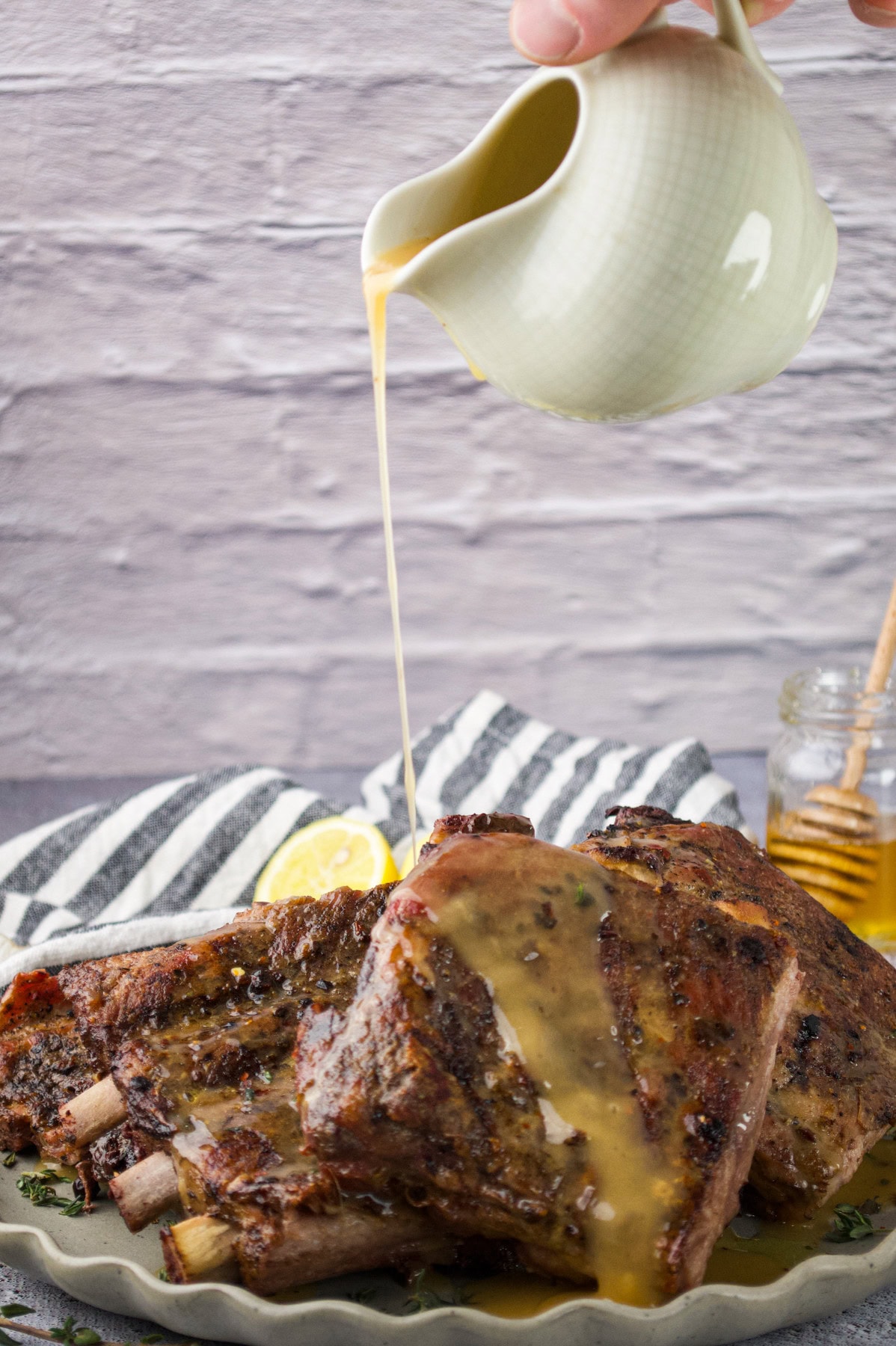A hand pours golden sauce from a small pitcher onto a plate of seasoned, grilled ribs. The ribs rest on a striped cloth, with a slice of lemon and a jar of honey visible in the background.