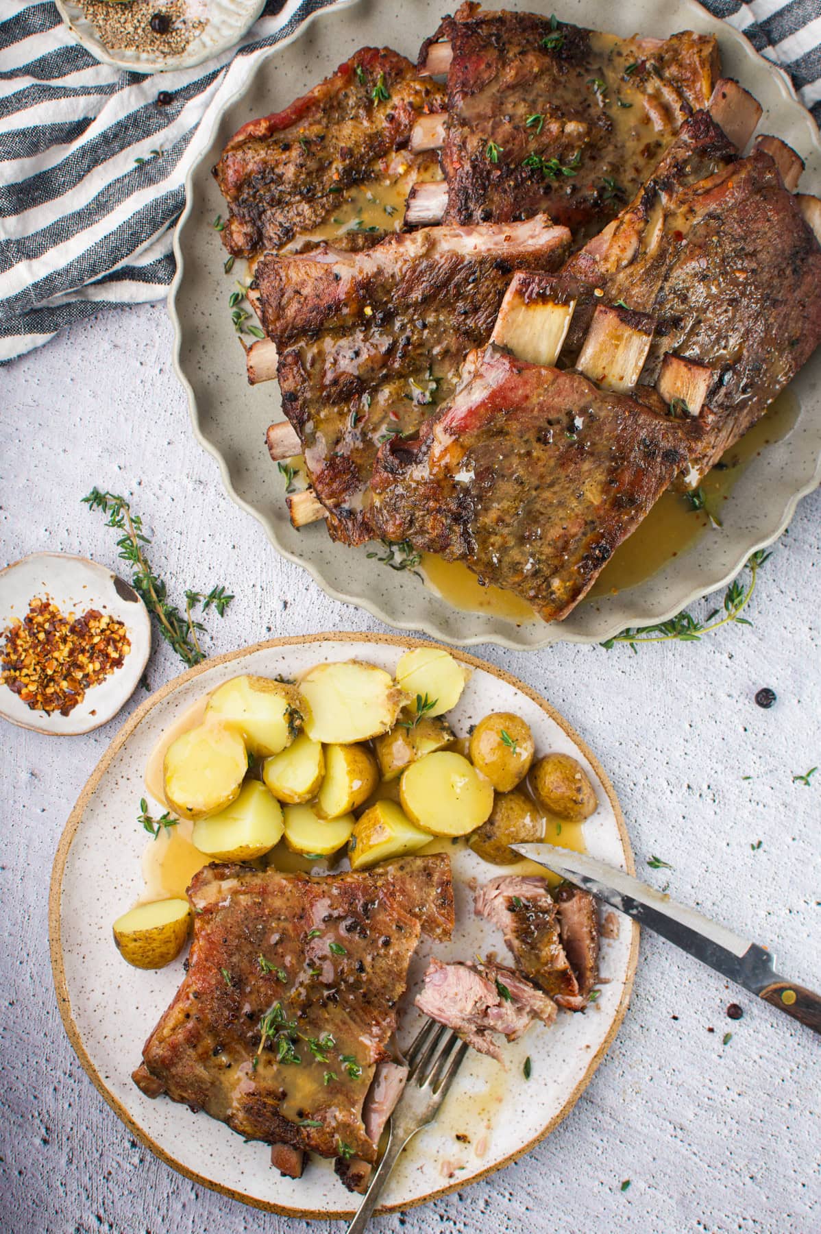 A plate of Juniper Berry Baby Back Ribs with herbs and sauce, next to a dish of sliced boiled potatoes. A fork holds a piece of rib on the smaller plate. A striped towel and seasonings are nearby on a light-colored surface.