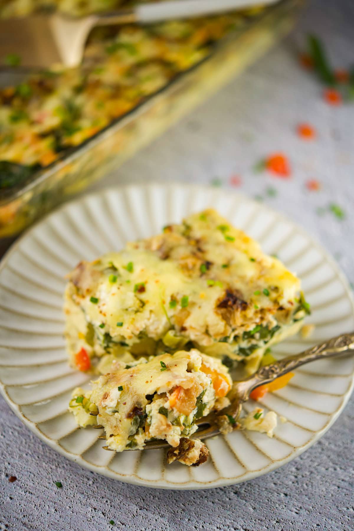 A close-up of a serving of vegetable casserole on a white plate with a forkful taken, showing a creamy mixture of vegetables and herbs. The casserole dish is visible in the background on a light surface.