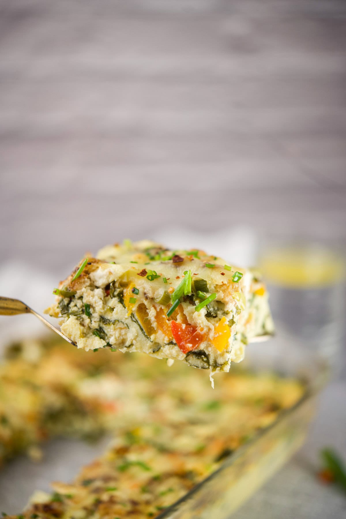 A close-up of a metal spatula holding a slice of vegetable casserole, topped with chopped herbs, with a blurred casserole dish and glass in the background.