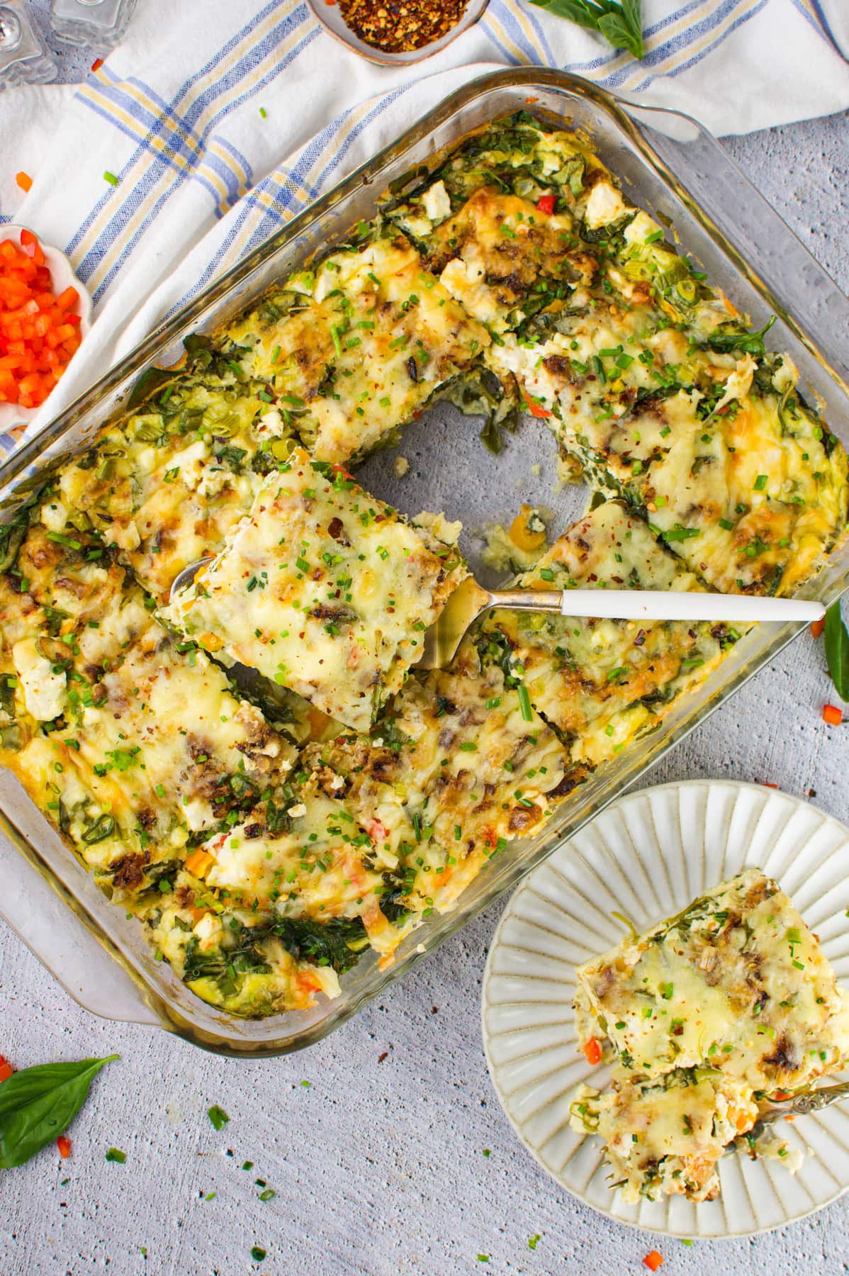 A glass baking dish filled with vegetable lasagna, one square piece being lifted out with a spatula. A serving of lasagna is plated nearby, with chopped vegetables and herbs scattered around.