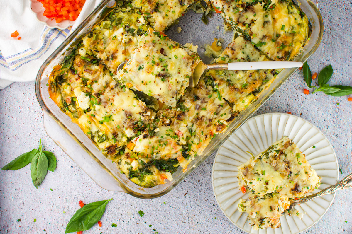 A Cottage Cheese Egg Bake with a slice being served onto a white plate. Fresh basil leaves and chopped vegetables are scattered around on a light gray surface.