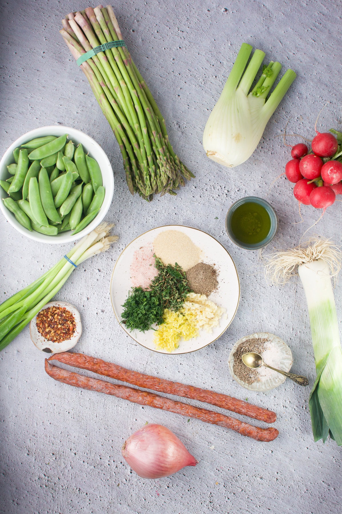 A top-down view showcases the ingredients for One-Sheet Pan Spring Roasted Vegetables (With Polish Sausage): asparagus, fennel, radishes, sugar snap peas, leek, shallot, sausages, spices, olive oil, and a plate of chopped herbs and garlic.