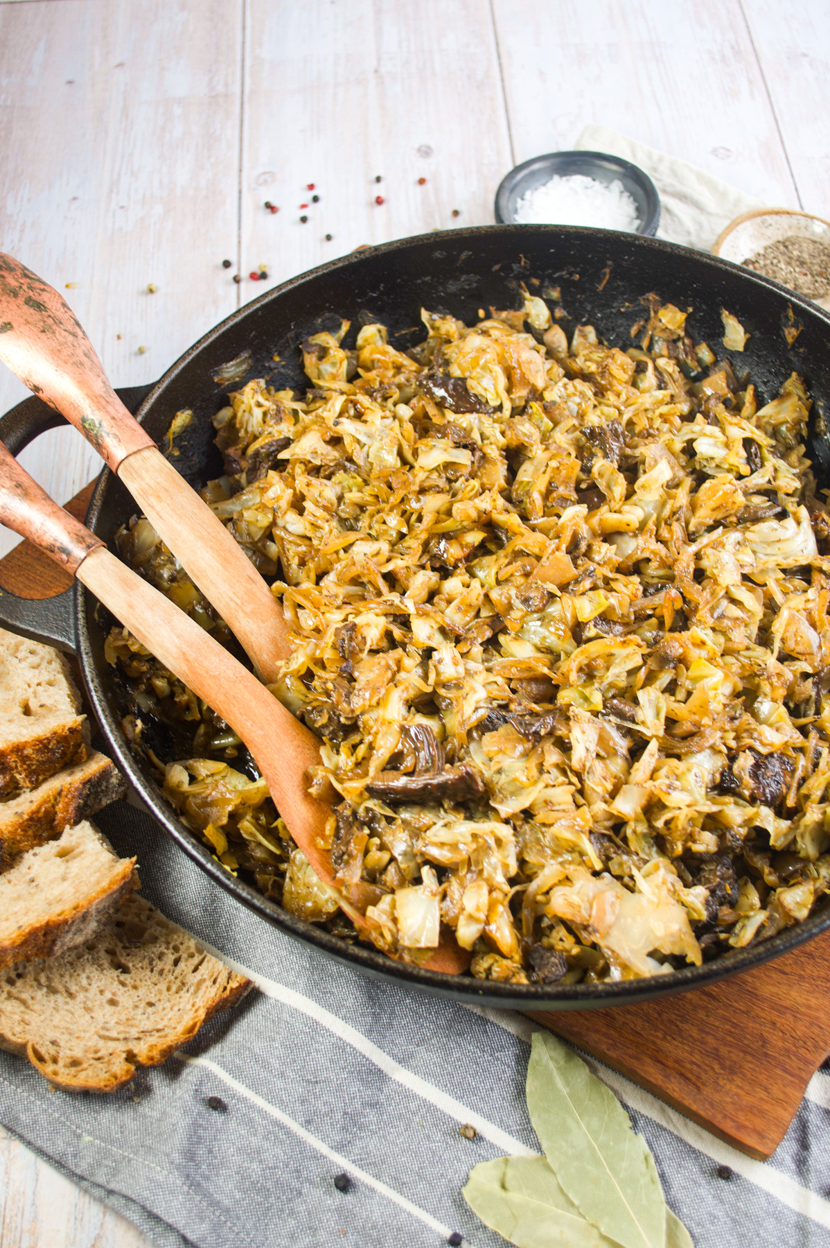 A skillet filled with Polish sautéed cabbage and wild mushrooms sits on a wooden board, with wooden utensils resting inside. Slices of bread, bay leaves, and small bowls of spices are nearby on a striped cloth.