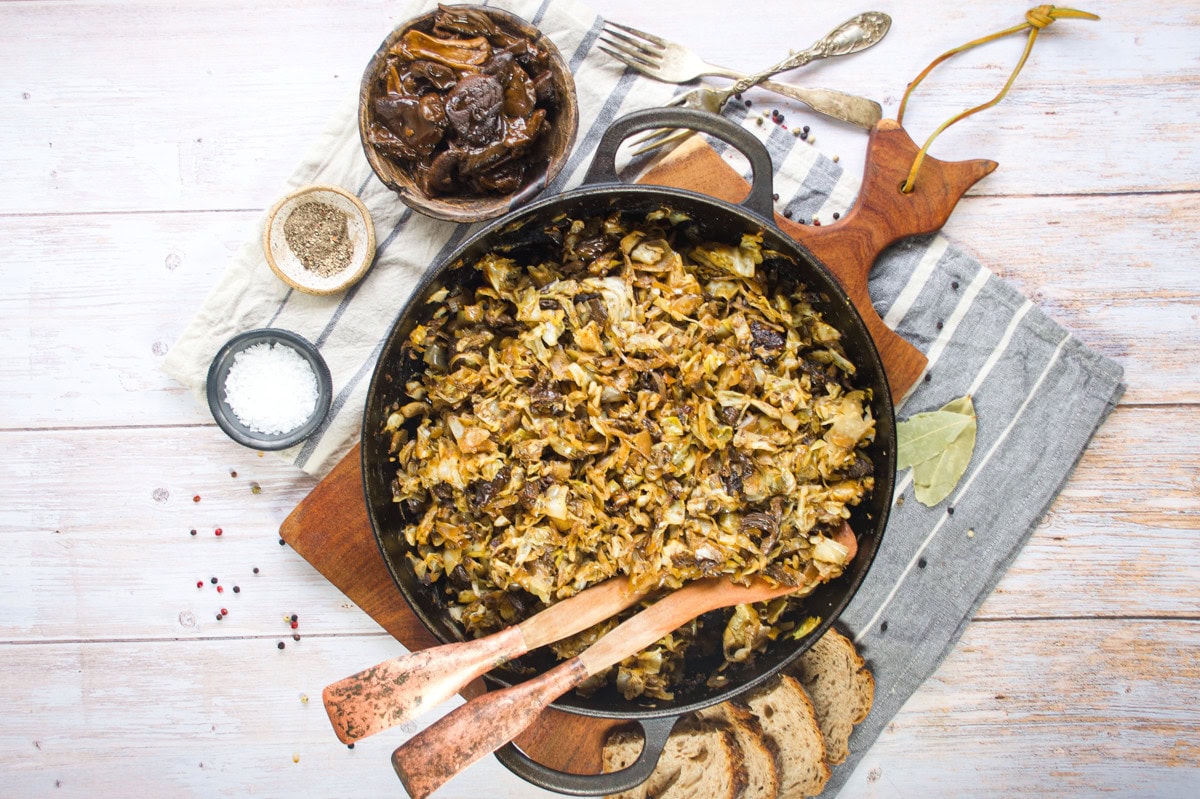 A skillet filled with braised cabbage and mushrooms sits on a wooden board. Slices of bread, bowls of salt and pepper, a small dish of mushrooms, and utensils surround the skillet on a striped cloth.