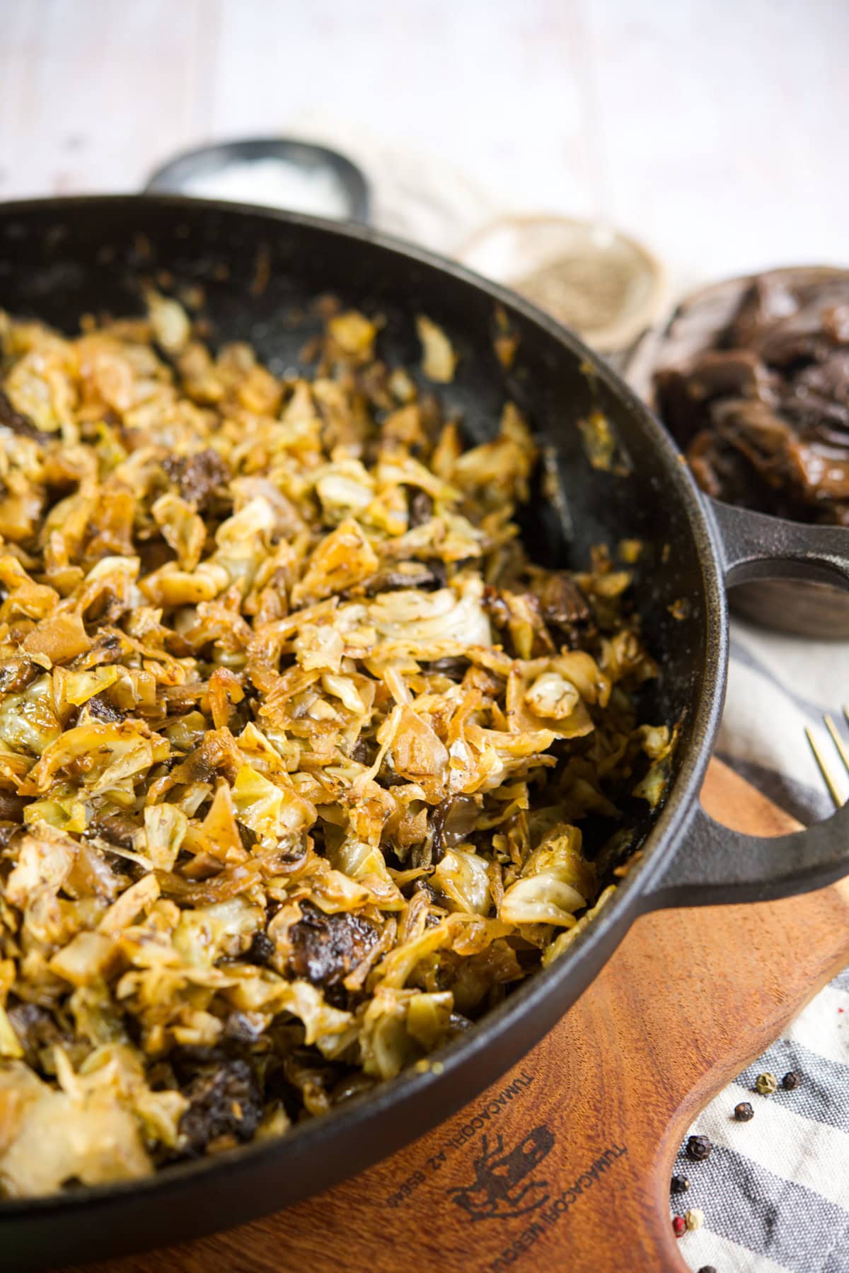 A cast iron skillet filled with saut&eacute;ed cabbage and mushrooms sits on a wooden board, with a bowl of mushrooms and a fork in the background. The dish appears golden-brown and freshly cooked.