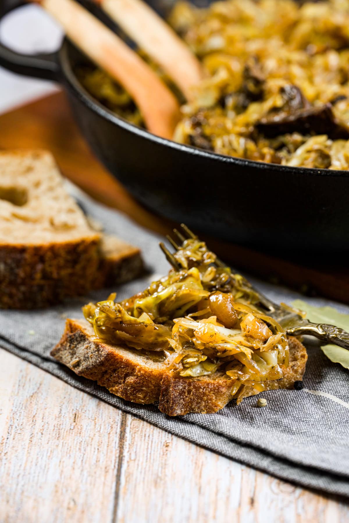 A close-up of saut&eacute;ed cabbage and mushrooms served on a slice of rustic bread, with more of the mixture in a skillet in the background and a fork resting on the bread, all on a wooden table.
