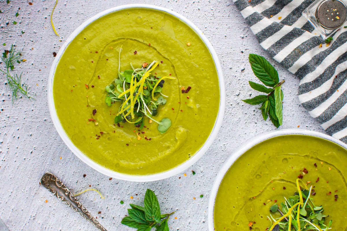 Two bowls of creamy green soup garnished with microgreens and yellow sprouts, placed on a light textured surface next to fresh mint leaves, a silver spoon, and a striped cloth napkin.