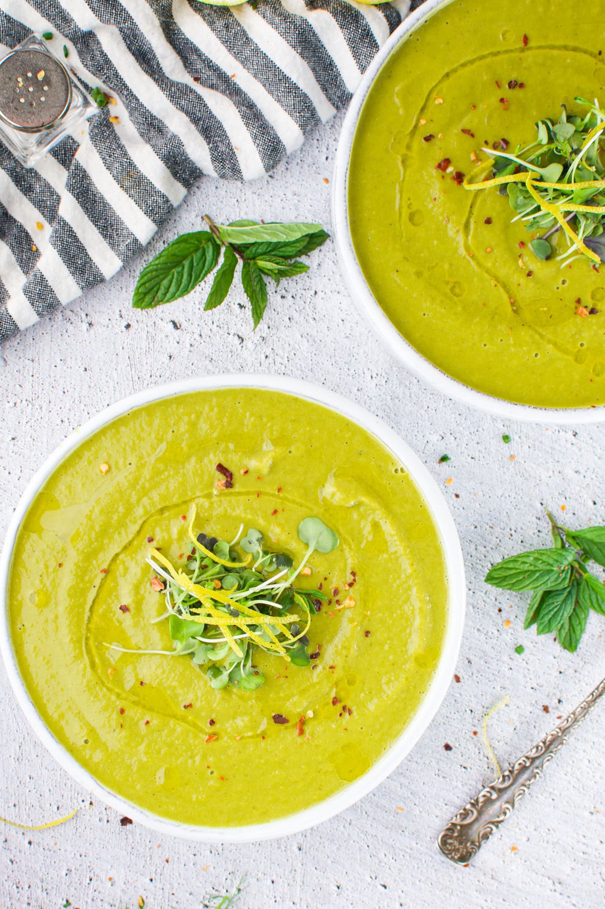 Two bowls of creamy green soup topped with microgreens and yellow zest sit on a white textured surface. A striped cloth, fresh mint, and a vintage spoon are nearby, adding a fresh and inviting presentation.