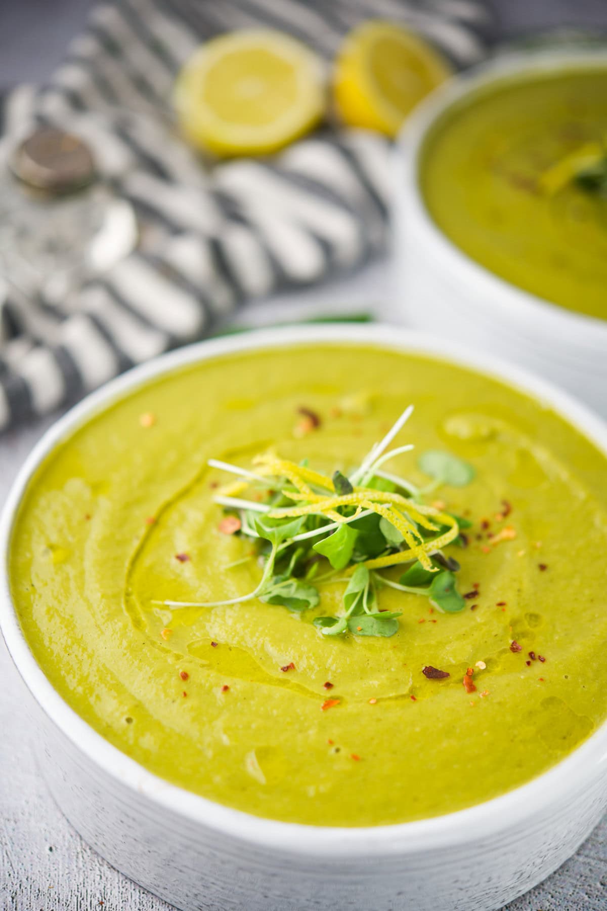 A bowl of creamy green soup garnished with microgreens, lemon zest, and red pepper flakes, with a striped cloth and lemon halves in the background.