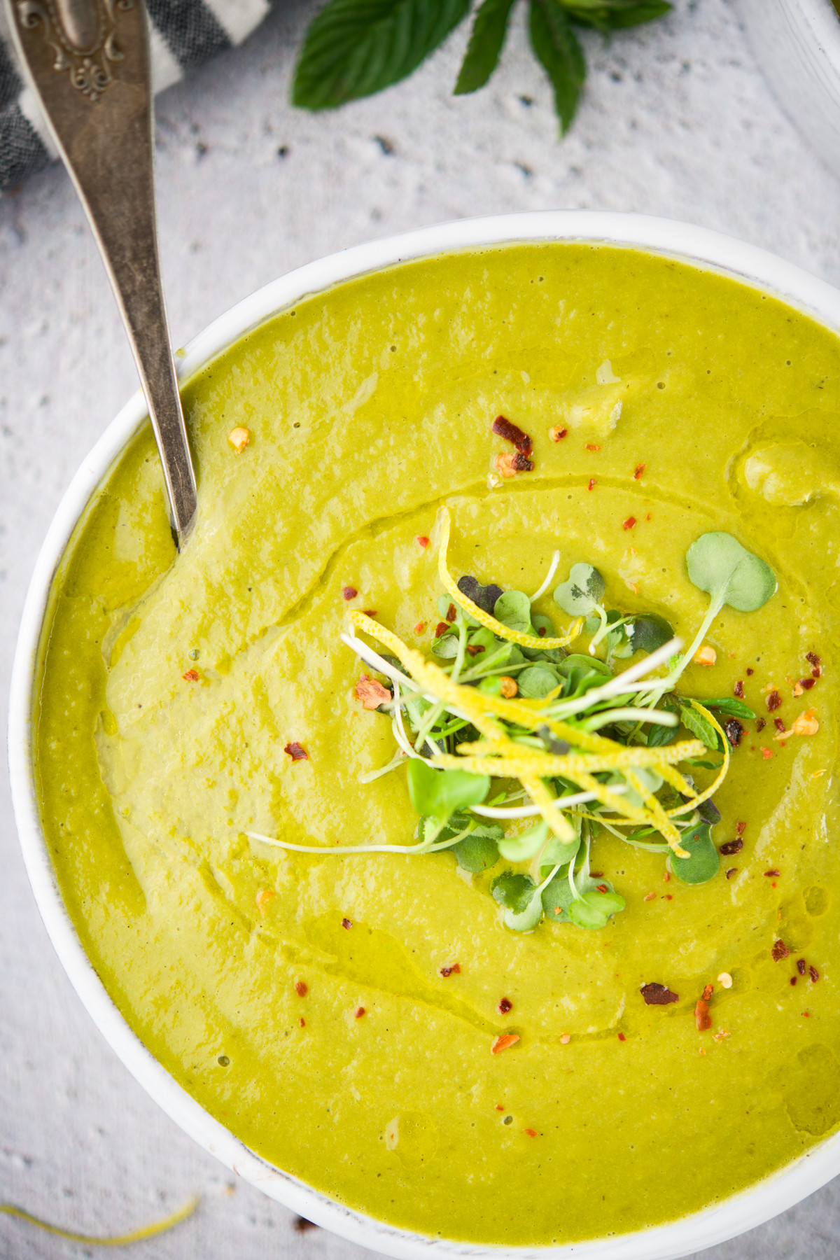 A bowl of Fennel, Lemon And Pea Soup garnished with microgreens and red pepper flakes, with a spoon resting inside the bowl on a light textured surface.