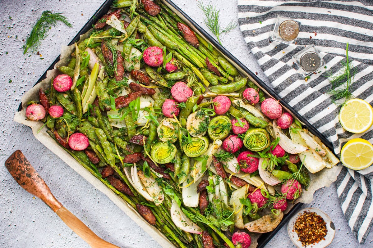 A baking tray lined with parchment paper holds roasted vegetables, including asparagus, radishes, artichokes, fennel, and sun-dried tomatoes, garnished with fresh herbs. Sliced lemons, spices, and utensils are nearby on a light surface.