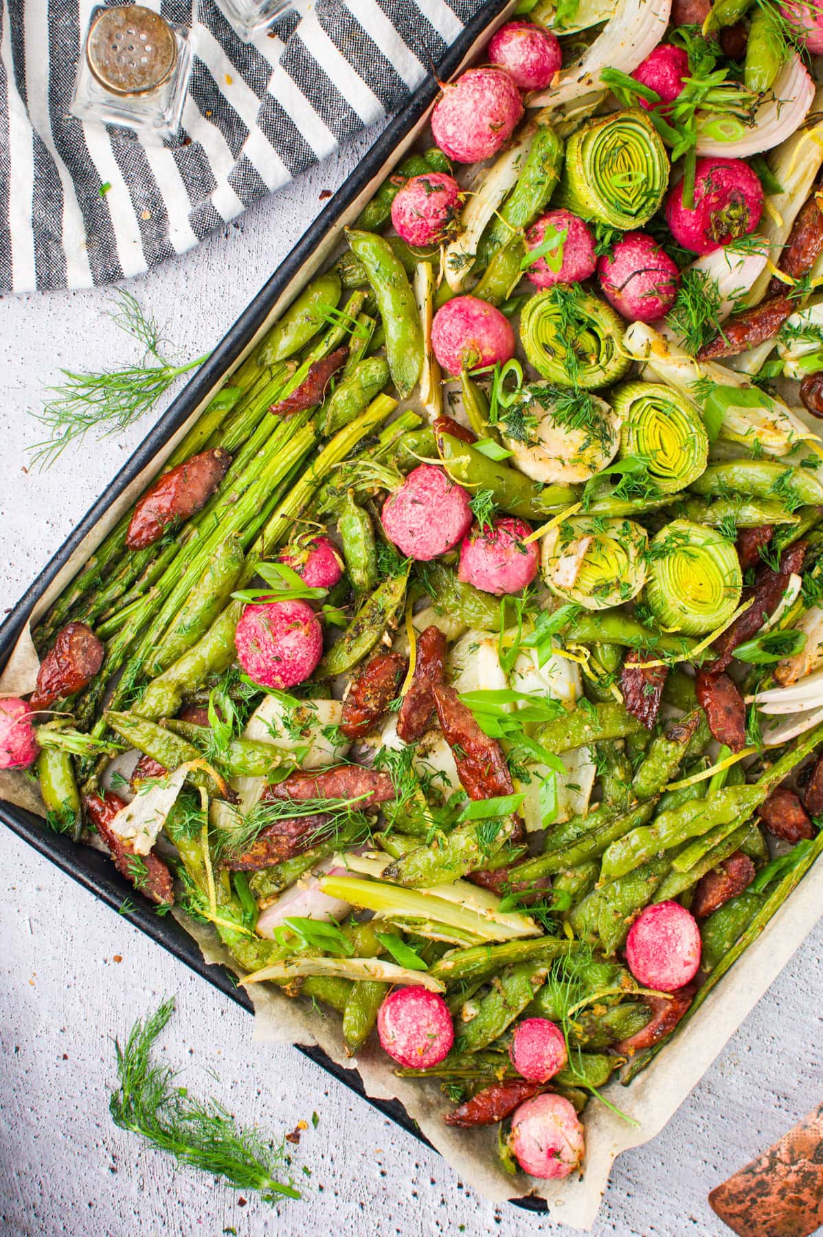 A baking tray filled with roasted green vegetables, radishes, leeks, green beans, asparagus, and sun-dried tomatoes, garnished with fresh dill and green onions, on a light surface next to a striped cloth.