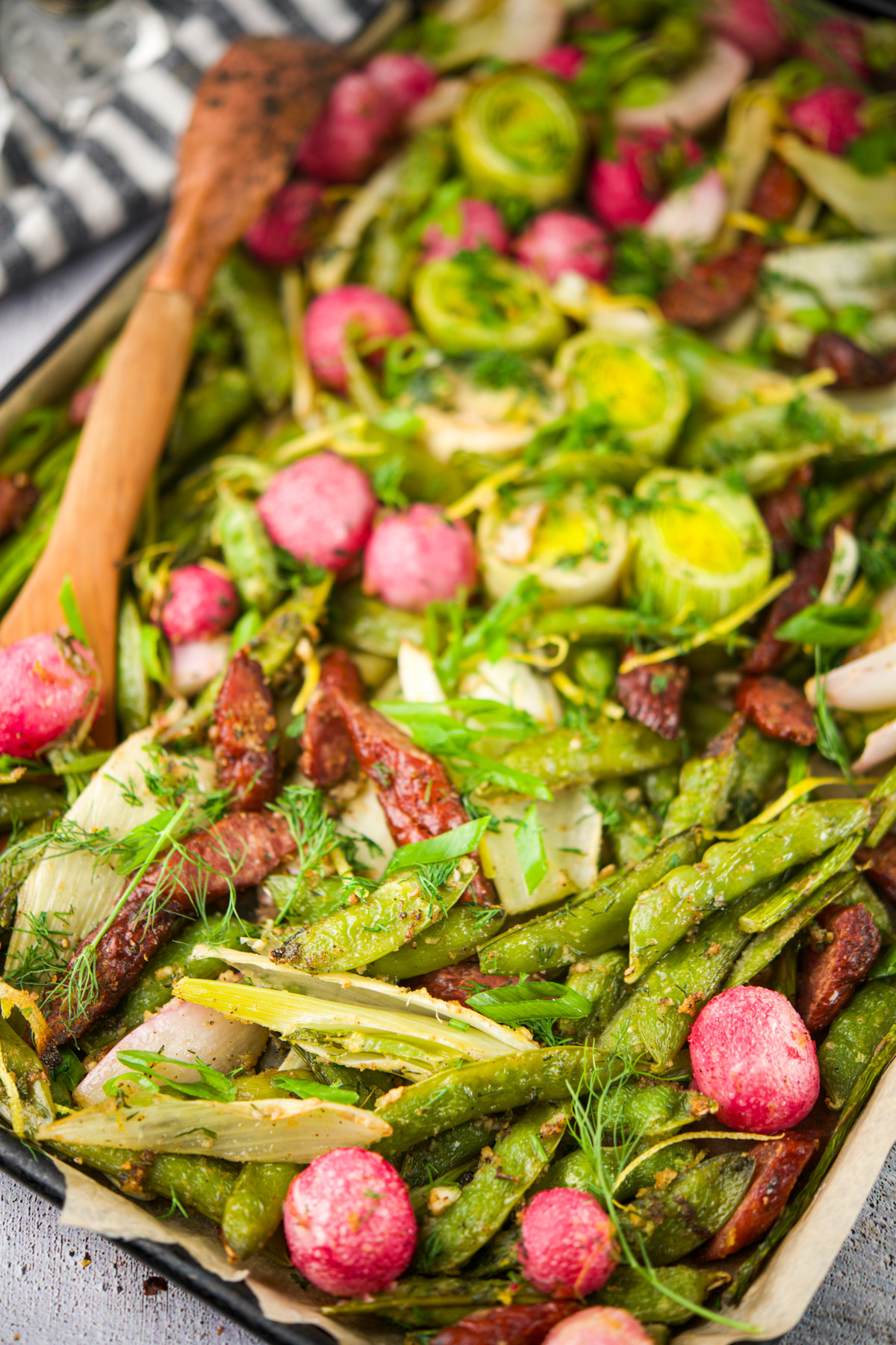 A tray of roasted vegetables including green beans, Brussels sprouts, radishes, and sun-dried tomatoes is topped with fresh herbs. A wooden spoon rests on the tray, ready for serving.