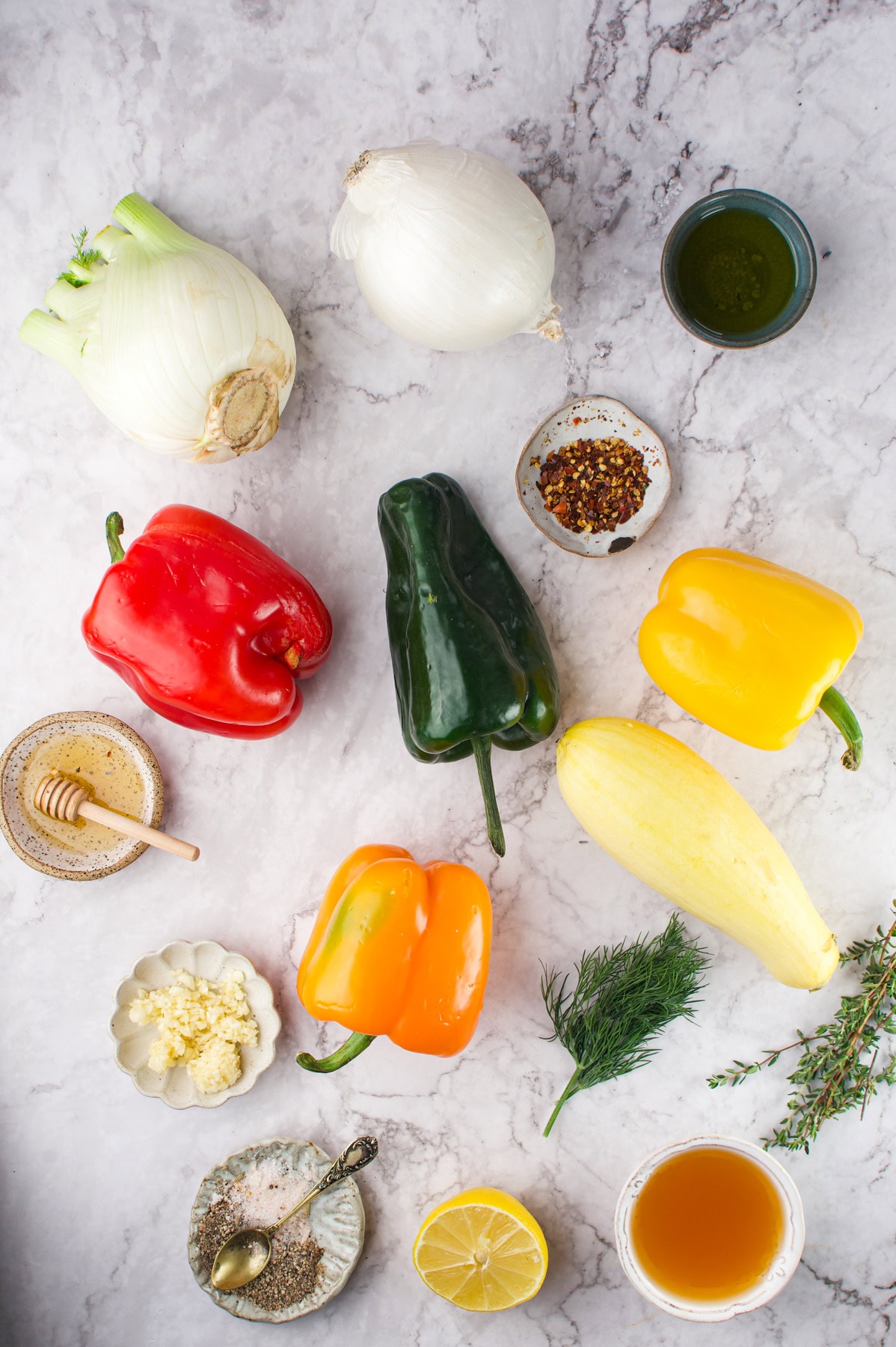 Colorful bell peppers, squash, fennel, onion, herbs, honey, minced garlic, lemon, olive oil, broth, and red pepper flakes arranged on a marble surface, viewed from above.