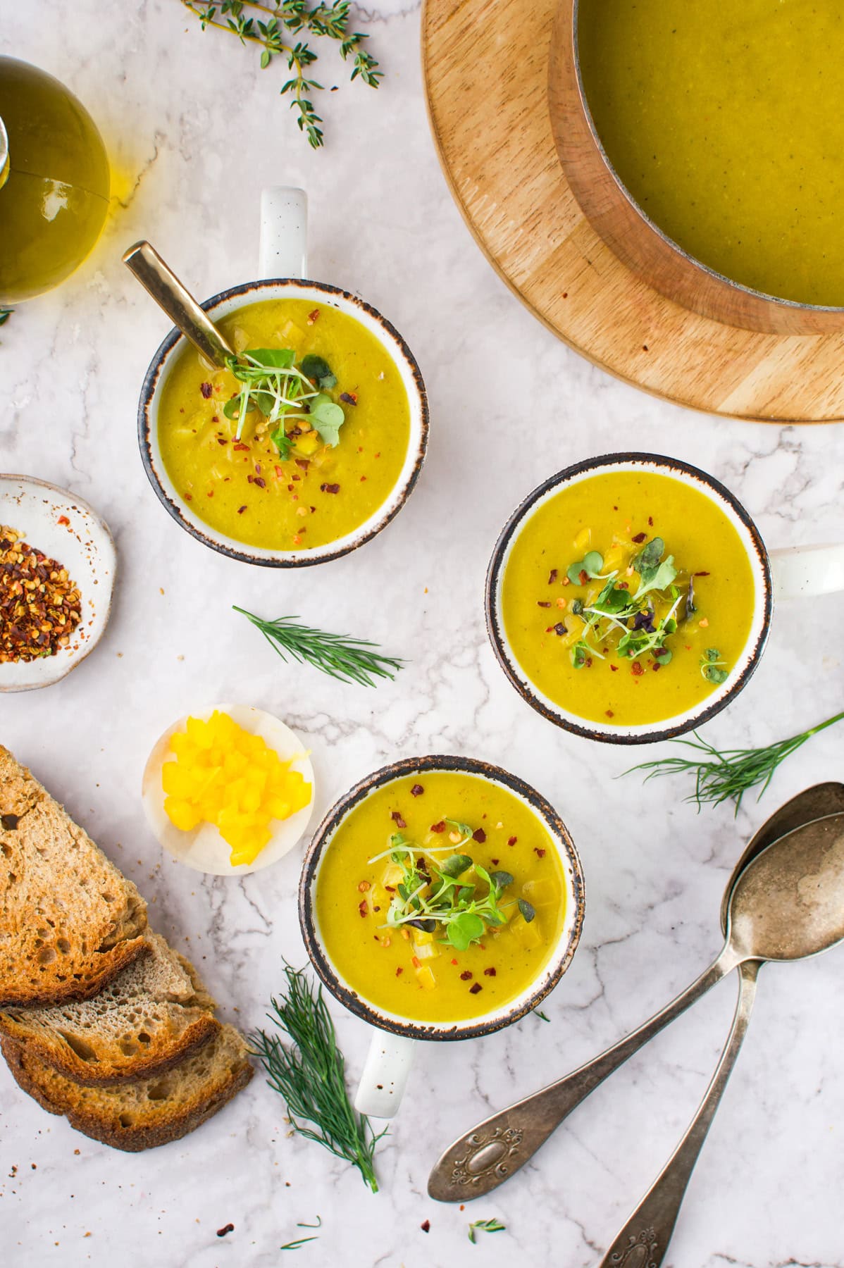 Three mugs of Creamy Fennel Yellow Squash Soup garnished with herbs, surrounded by slices of bread, fresh dill, a bowl of yellow pickles, red pepper flakes, two spoons, and a pot of soup on a marble surface.