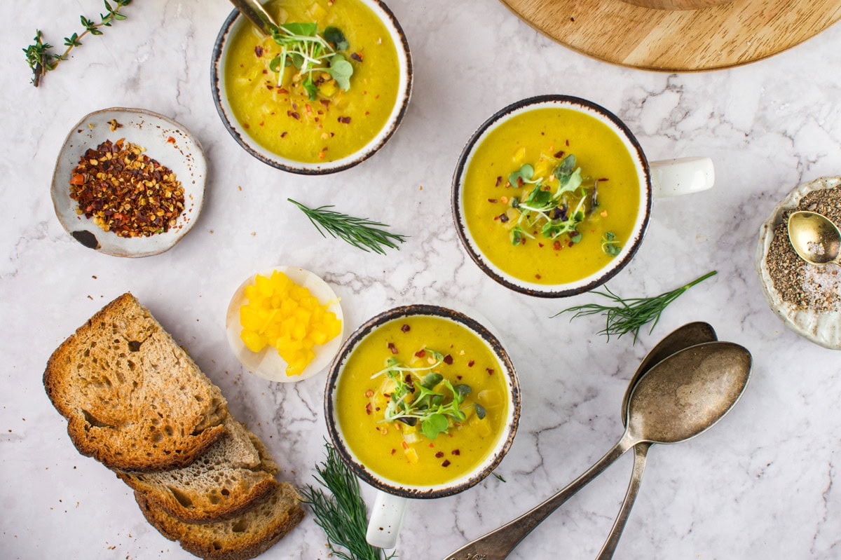 Three bowls of yellow soup garnished with microgreens sit on a marble surface, surrounded by slices of toasted bread, yellow diced vegetables, herbs, red pepper flakes, spoons, and a bowl of seeds.