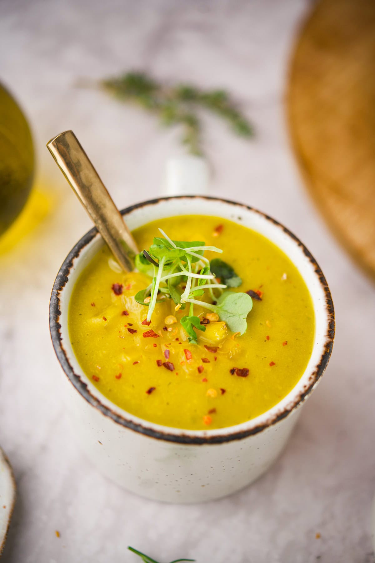 A white ceramic mug filled with creamy yellow soup, garnished with microgreens, red pepper flakes, and a gold spoon. The mug sits on a light marble surface with blurred herbs in the background.