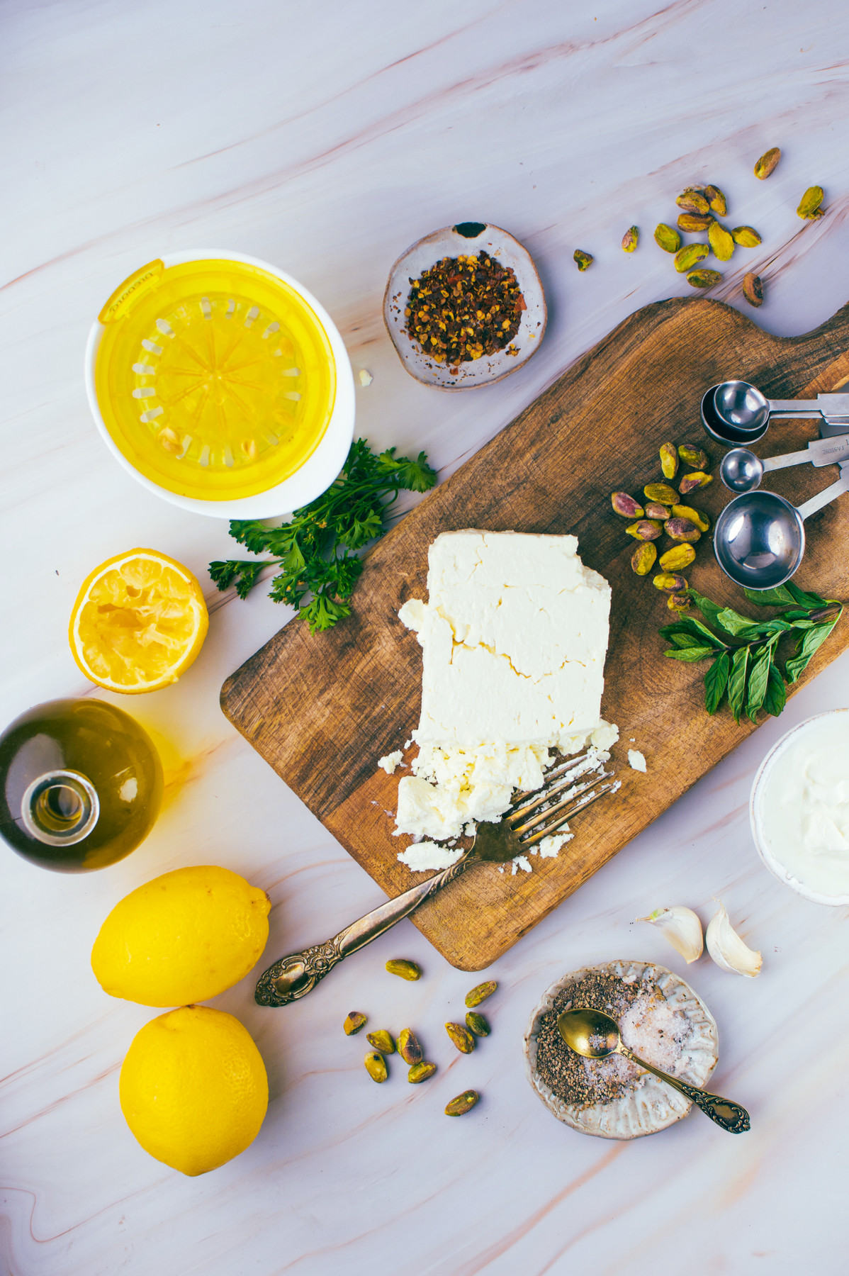 Wood board with feta cheese, a fork, pistachios, and herbs. Around it are lemons, olive oil, a citrus juicer, spices, garlic, metal spoons, and a bowl of yogurt on a white surface. Ingredients arranged for cooking.
