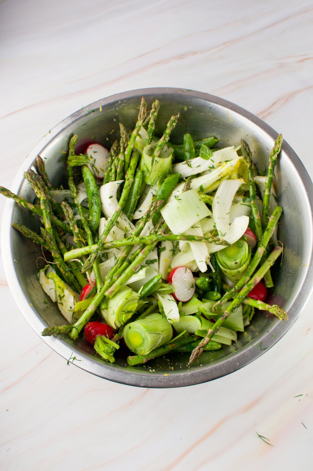 A stainless steel bowl filled with fresh vegetables, including asparagus, leeks, radishes, and herbs, sits on a light marble surface, ready for preparation or cooking.