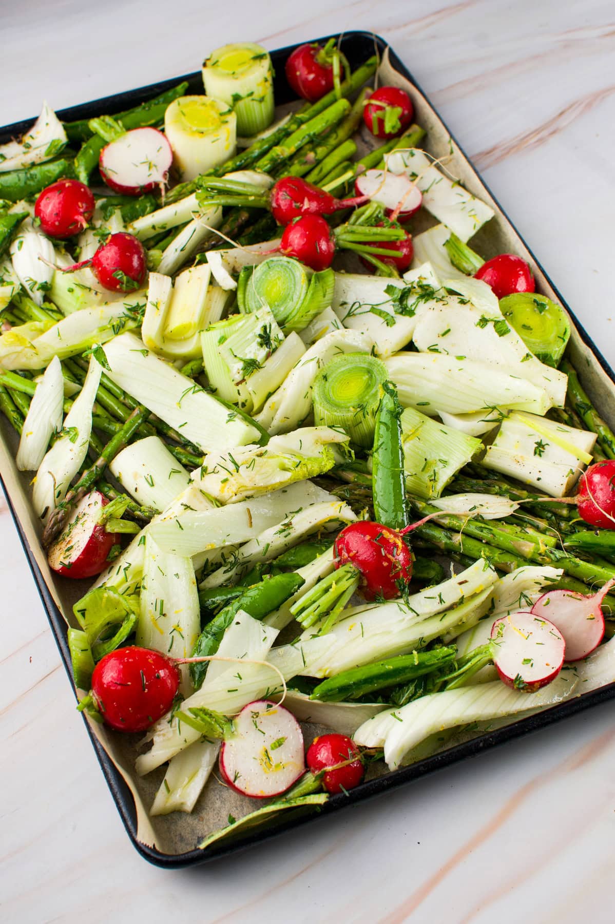 A baking tray filled with Roasted Spring Vegetables, including sliced leeks, radishes, asparagus, and fresh herbs, arranged on parchment paper, ready for roasting.