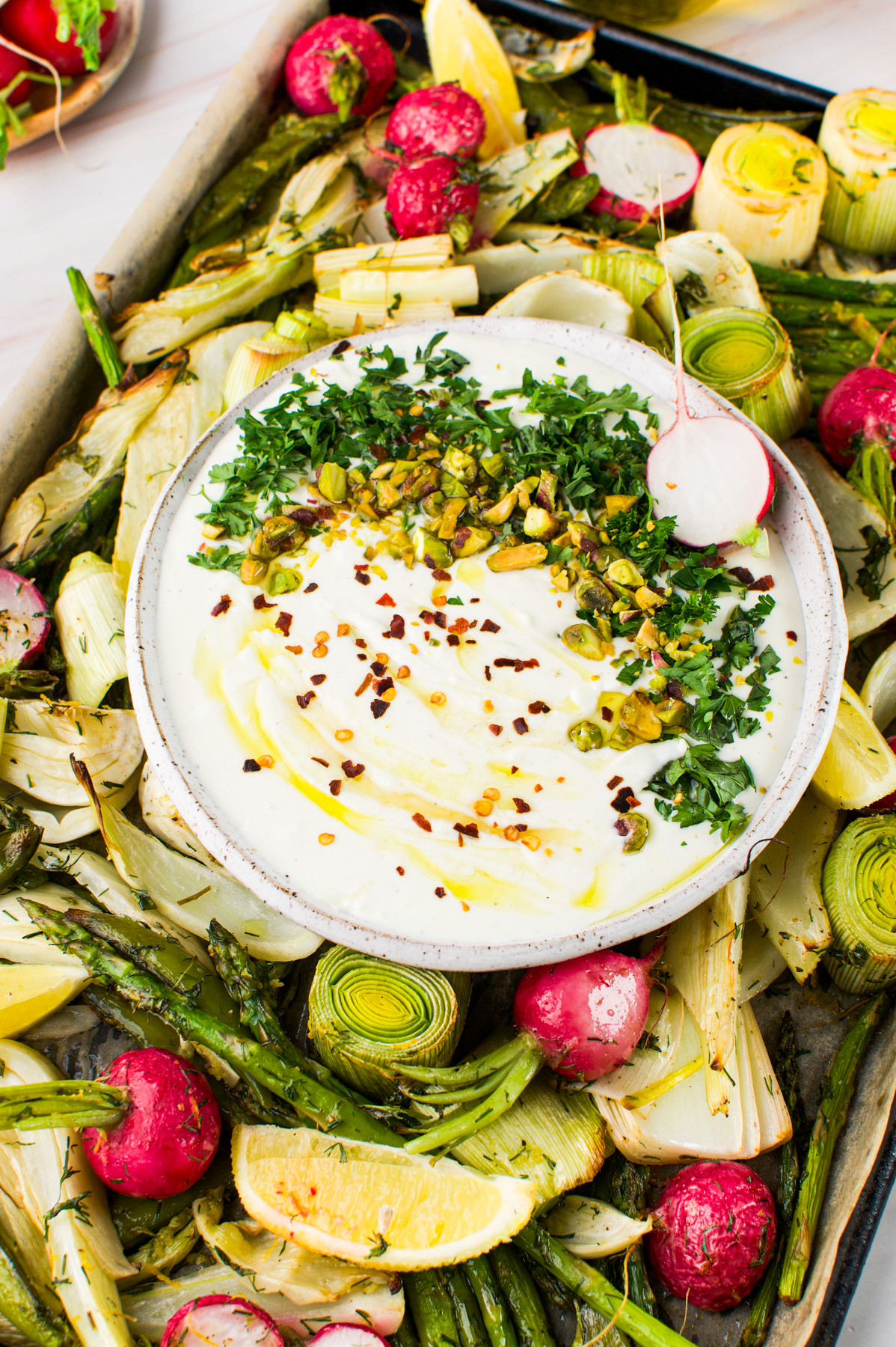 A bowl of creamy dip topped with herbs, nuts, and chili flakes is surrounded by roasted vegetables like asparagus, radishes, leeks, and lemon wedges on a tray.