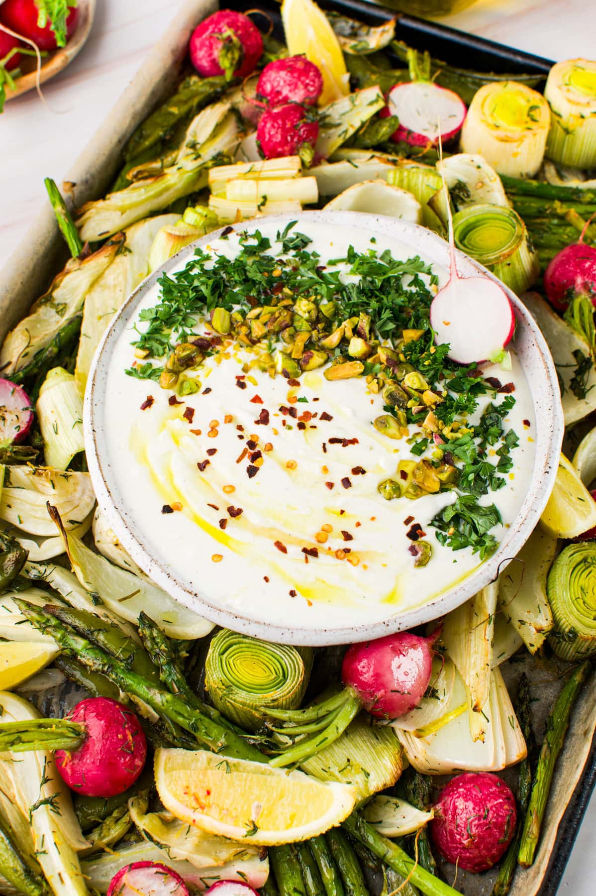 A bowl of creamy dip topped with chopped pistachios, parsley, chili flakes, and olive oil sits on a tray surrounded by roasted vegetables, including radishes, asparagus, leeks, artichoke, and lemon wedges.